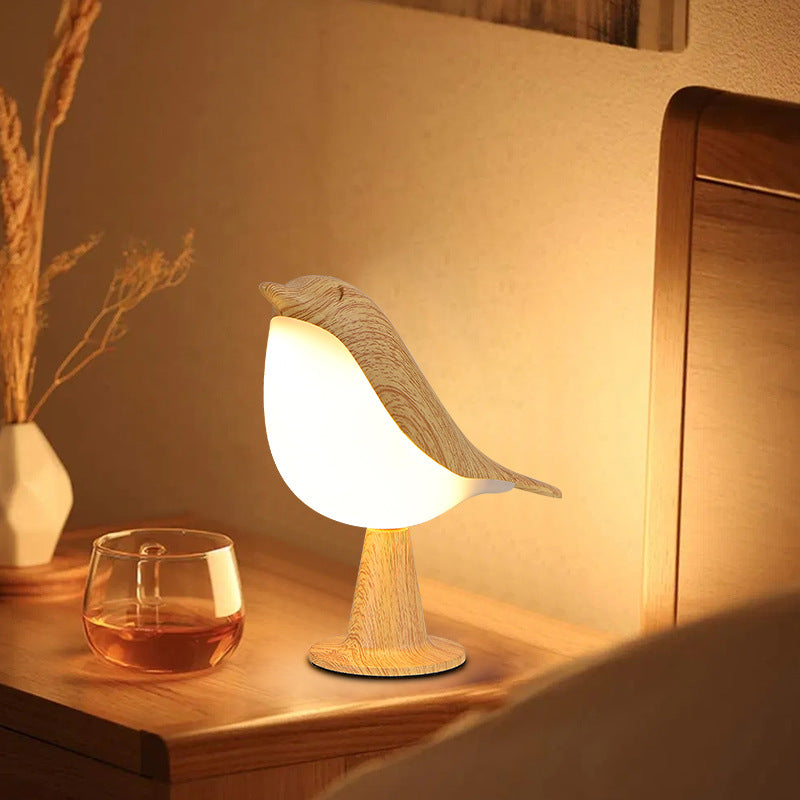 Bird-shaped night light on a wooden surface with a glass of liquid and a vase in the background.