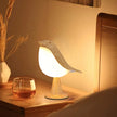 Bird-shaped night light on a wooden surface with a glass of liquid and a vase in the background.