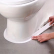 Person applying a white strip to the base of a toilet on a gray floor.