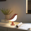 Bird-shaped table lamp on a desk with a plant and newspaper in the background